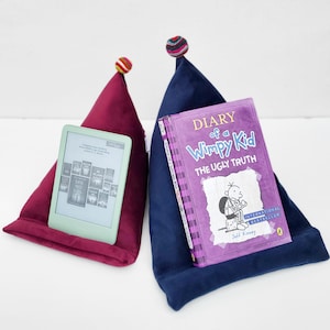 May include: Two plush book holders, one burgundy and one navy blue, each with a pom-pom on top. The burgundy holder has an e-reader, and the navy holder has a book titled "Diary of a Wimpy Kid: The Ugly Truth."