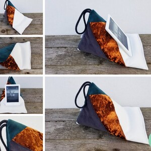 Puede incluir: Una almohada de tela blanca, gris, verde y naranja con cierre de cordón. La almohada tiene forma de triángulo y está diseñada para sostener una tableta o un libro.