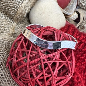 May include: A silver-colored bracelet with a hammered texture rests on a red woven sphere. The bracelet has a curved design. The background includes a red knitted fabric, burlap, and decorative elements.