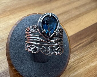 Puede incluir: Un anillo de plata con una gran piedra preciosa azul oscuro en forma de pera. El anillo tiene un diseño detallado y en capas con un patrón floral en la base. El anillo se exhibe en un soporte gris.