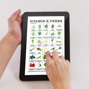 May include: A person holding a black tablet displaying a Vitamin K Foods chart. The chart lists various vegetables and fruits with their Vitamin K content per 1000 units. The text "Vitamin K Foods" is at the top.