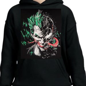 May include: Black hooded sweatshirt with a graphic print of the Joker and Venom. The graphic shows the Joker's face with green hair and a wide smile, and Venom's face with a black and red tongue. The text "HA HA HA" is printed in green on the sweatshirt.
