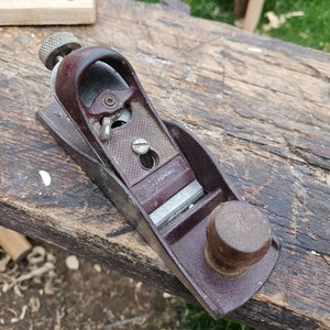 Puede incluir: Un cepillo de mano de madera vintage con un cuerpo de metal marrón y un mango de madera. El cepillo tiene una cuchilla de metal y una pequeña perilla en la parte superior.
