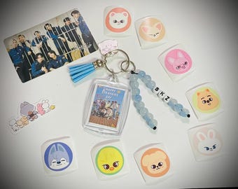 May include: An assortment of stickers, a keyring, and a photo card. The keyring has a blue tassel and a clear case with a photo. The stickers feature cartoon animal faces. The photo card shows a group of people.