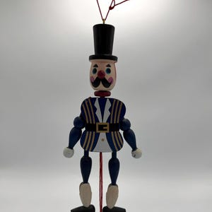 Puede incluir: Soldado de juguete de madera con cuerda, con sombrero de copa negro, cara pintada con bigote y uniforme azul y dorado. El juguete tiene extremidades articuladas y una cuerda roja. El cinturón del soldado tiene una hebilla dorada.