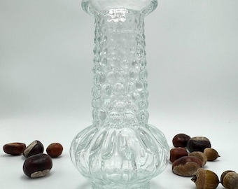 May include: A clear glass vase with a textured, bubble-like pattern on the body. The vase has a flared top and a rounded base. Scattered around the vase are several brown chestnuts and acorns, suggesting a fall theme.