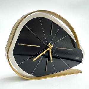 May include: A vintage black and gold-tone clock with a unique, asymmetrical design. The clock face is black with white lines radiating from the center. The hands and frame are gold-tone. The clock has a curved, arched handle.