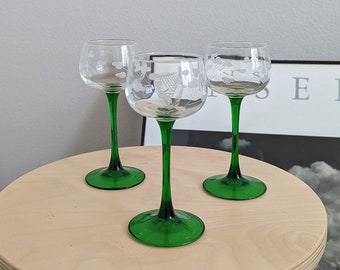 May include: Three clear glass wine glasses with etched floral designs and green stems on a light wood table.