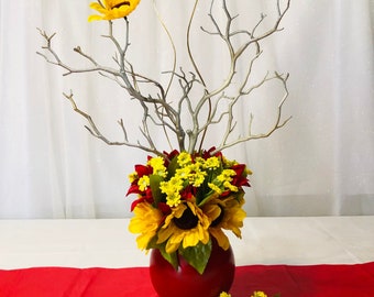 Puede incluir: Arreglo floral vibrante en un jarrón rojo. El arreglo presenta girasoles, flores amarillas y detalles rojos, con ramas y alambres dorados. Se encuentra sobre un camino de mesa rojo.