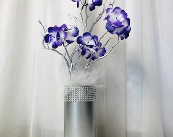 Puede incluir: Un jarrón plateado con un diseño brillante de diamantes está lleno de un arreglo floral blanco y morado. El jarrón está sobre una mesa blanca.