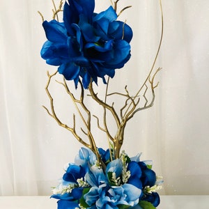 Puede incluir: Un centro de mesa de ramas doradas con flores artificiales azules y azul claro en un jarr&oacute;n dorado.