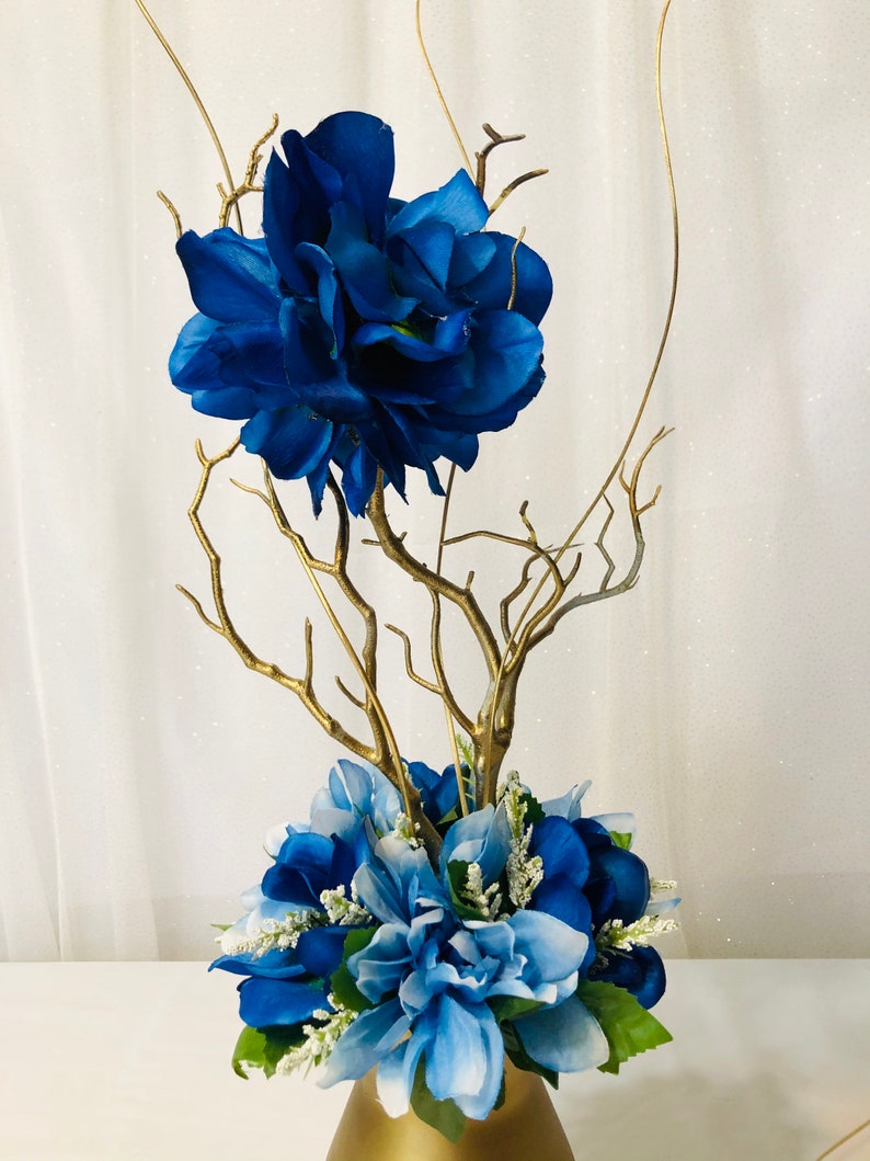 Puede incluir: Un centro de mesa de ramas doradas con flores artificiales azules y azul claro en un jarr&oacute;n dorado.