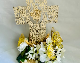 May include: A wooden cross with the words "Padre Nuestro" and the Lord's Prayer in Spanish, decorated with white flowers and gold butterflies on a wooden base.