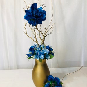 Puede incluir: Un jarr&oacute;n dorado con flores de seda azules y azul claro dispuestas en ramas doradas. El jarr&oacute;n est&aacute; sobre un mantel blanco.