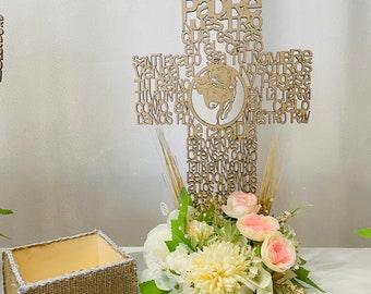 May include: A wooden cutout of the Lord's Prayer in Spanish, "Padre Nuestro", with a floral arrangement in a burlap-wrapped box. The flowers are white and pink, with green leaves. The arrangement is on a mirrored surface.