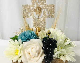 May include: A burlap flower arrangement with white and blue flowers, black grapes, and a wooden cross with the text "Padre Nuestro" in Spanish. The arrangement is in a burlap container with a gold ribbon and the word "Communion" on it.