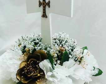 May include: A white wooden cross with a gold-toned design sits atop a floral arrangement. The arrangement features white carnations, white baby's breath, and a single brown rose with gold accents.