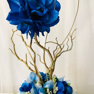 Puede incluir: Un centro de mesa floral azul y dorado con una gran flor azul en la parte superior de un arreglo de ramas doradas. El arreglo est&aacute; rodeado de flores azules m&aacute;s peque&ntilde;as.