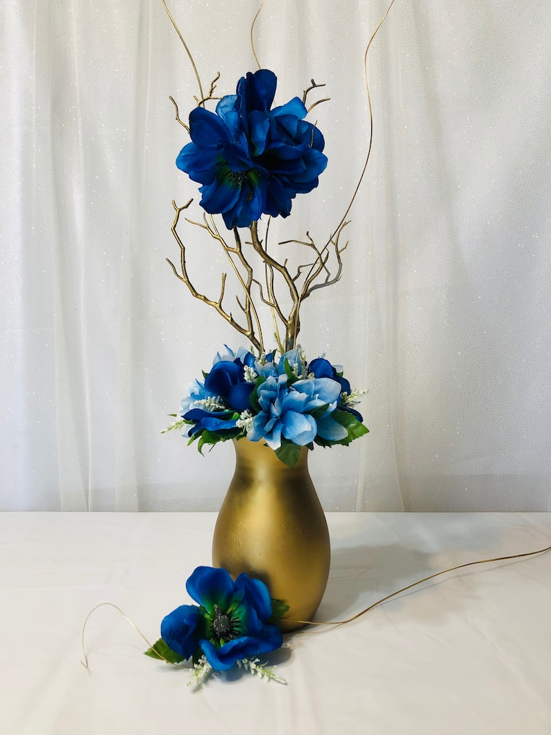 Puede incluir: Un jarr&oacute;n dorado con flores azules y ramas doradas. El jarr&oacute;n est&aacute; sobre un mantel blanco.