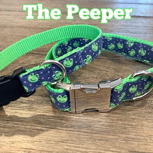 May include: Two dog collars. One is fastened with a black plastic buckle, the other with a silver metal clasp. Both collars have a green nylon strap and a blue fabric with green frog faces. The text "The Peeper" is at the top.