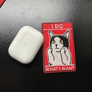 May include: A white wireless earbud case next to a red patch with the text "I DO WHAT I WANT" and a cat illustration. The patch has a black and white cat with its middle fingers up.