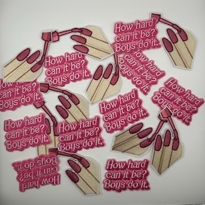 May include: Embroidered patches with a hand design and the text "How hard can it be? Boys do it." The patches feature a beige hand with pink nails and a pink text box. The patches are scattered on a white surface.