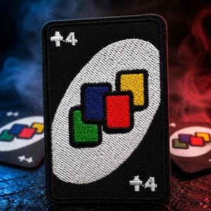 May include: A rectangular embroidered patch featuring a black background with a white oval in the center. Inside the oval are four small, overlapping squares in green, blue, red, and yellow. The text +4 is embroidered in white in the top left and bottom right corners.