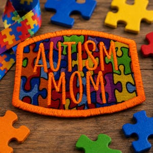 May include: A rectangular embroidered patch with an orange border featuring a colorful puzzle piece pattern in red, blue, green, and yellow. The text AUTISM MOM is stitched in orange thread across the center of the patch, which is surrounded by loose puzzle pieces.