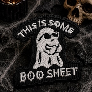 May include: A black embroidered patch featuring a white ghost wearing sunglasses. The ghost is depicted with a hand near its chin. The text "THIS IS SOME" is embroidered in white above the ghost, and "BOO SHEET" is embroidered in white below.