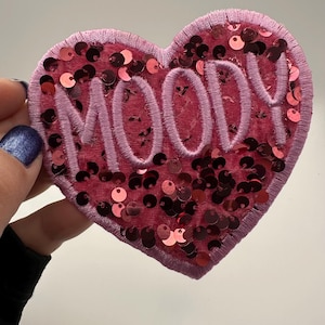 May include: A heart-shaped patch with the word "MOODY" embroidered in light pink thread. The patch is made of pink fabric and embellished with red sequins. The patch has a light pink border.