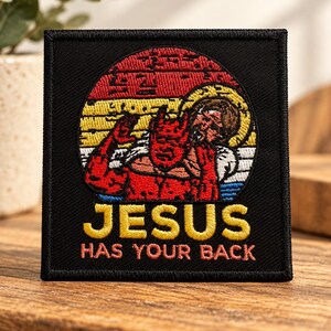 May include: A square black embroidered patch featuring a red devil and a figure with a halo against a sunset background of red, yellow, and white stripes. The text "JESUS" in yellow and "HAS YOUR BACK" in pink is stitched below the graphic.