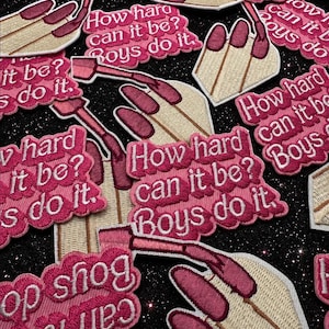 May include: A collection of embroidered patches featuring a hand with long dark pink nails holding a lipstick tube, paired with a pink scalloped speech bubble containing the white embroidered text: How hard can it be? Boys do it.