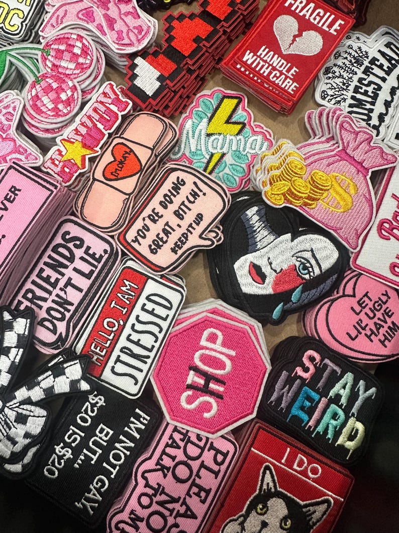 May include: A collection of embroidered patches in various shapes and colors. Patches include phrases like "Stay Weird," "Hello, I am Stressed," and "Shop." Other designs feature a crying face, a heart, and a cat.