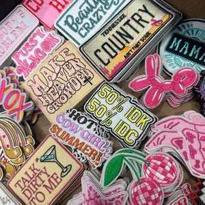 May include: A collection of embroidered patches with various designs and text. The patches feature phrases like "Beautiful Crazy," "Country," and "Talk Dirty to Me." Colors include pink, white, yellow, and green. The patches are likely for clothing or accessories.