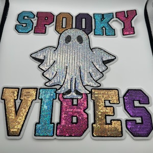 May include: A white sequined ghost patch with the words "SPOOKY VIBES" in colorful sequined letters. The letters are in gold, teal, pink, and purple. The ghost is silver and has a friendly face.