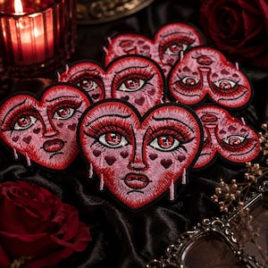 May include: A collection of heart-shaped embroidered patches featuring a stylized face with large eyes, long eyelashes, and a small mouth. The design incorporates shades of red and pink with small heart accents and vertical drip details.