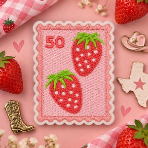 May include: A rectangular embroidered patch with a scalloped edge, featuring a light pink background, two red strawberries with green leaves, and the number 50 in red. The patch is surrounded by fresh strawberries, a small cowboy boot pin, a hat pin, and a Texas-shaped pin.
