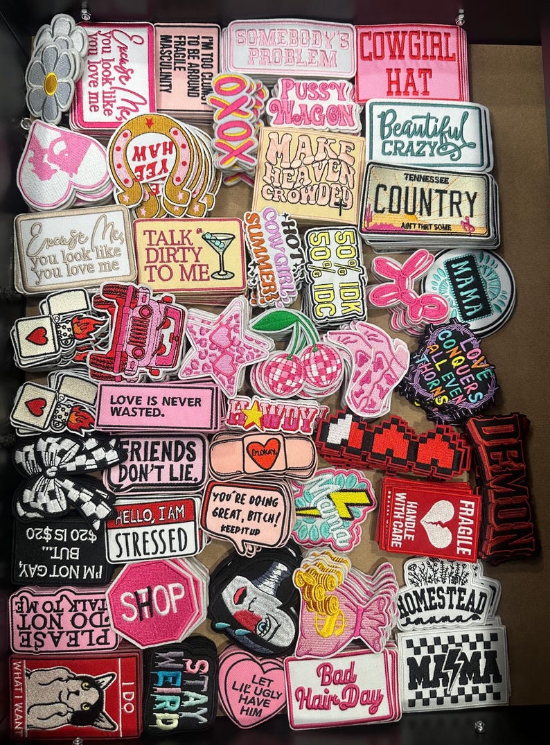 May include: A collection of embroidered patches in various shapes and colors. The patches feature text like "Cowgirl Hat," "Friends Don't Lie," and "Bad Hair Day." Other patches display images of a martini glass, cherries, and a cat.