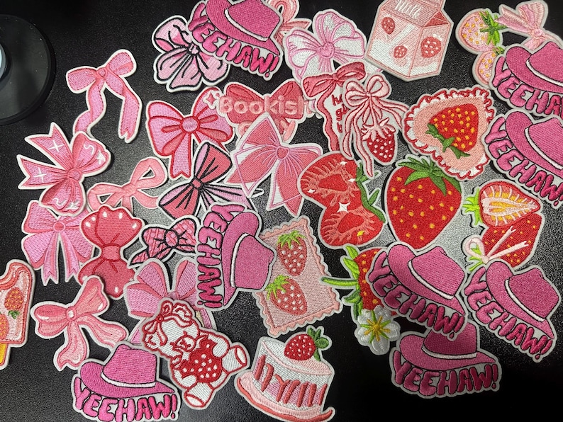 May include: A collection of pink embroidered patches featuring bows, strawberries, cowboy hats with "YEEHAW!" text, and a strawberry cake. The patches vary in size and design, with a white border.