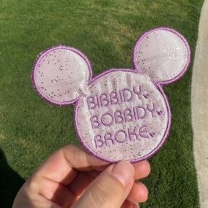 May include: A light purple, glittery, circular patch shaped like a mouse head with two rounded ears. The patch features dark purple embroidered text that reads "BIBBIDY BOBBIDY BROKE." The edges are finished with a dark purple stitched border.