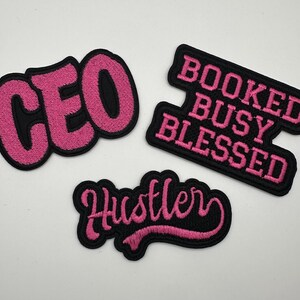 May include: Three embroidered patches with black borders and pink text. The patches read "CEO", "BOOKED BUSY BLESSED", and "Hustler". The patches are likely for clothing or accessories.