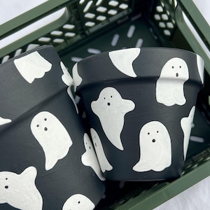 May include: Two black terracotta flower pots with white ghost illustrations. The pots are in a green plastic crate.