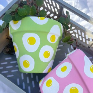 May include: Two painted terracotta flower pots, one green with white egg designs and one pink with white egg designs.