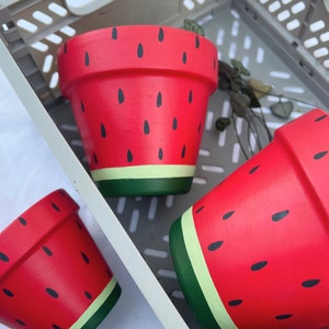 May include: Three terracotta flower pots painted to look like watermelons. The pots are red with black seeds and a green stripe around the top.