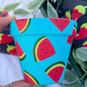 May include: A turquoise terracotta planter with a watermelon pattern. The planter is painted with red, green, and black watermelons on a light blue background.