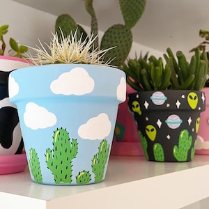 May include: Five different hand-painted terracotta flower pots. The pots are decorated with various designs, including clouds, cacti, and alien-themed patterns. The pots are all different sizes and shapes.