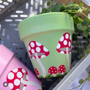 May include: Two painted terracotta flower pots with mushroom designs. The larger pot is green with white and red mushrooms. The smaller pot is pink with a white and red mushroom.