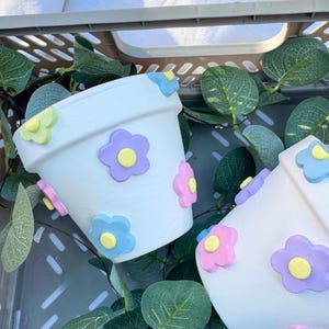 May include: White ceramic flower pots decorated with colourful, raised flower appliques in shades of pink, purple, yellow, and blue. The pots are set in a grey plastic crate with green leafy vines.