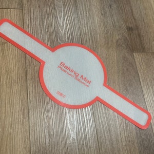 May include: A red-edged, white silicone baking mat with the words "Baking Mat Platinum Silicone" printed on it. The mat has a circular center and two rectangular extensions.