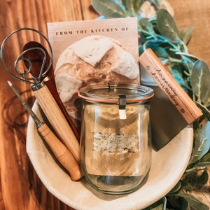 May include: A white bowl filled with baking supplies, including a wooden dough scraper, a wooden rolling pin, a dough whisk, a glass jar with a label that says "weck", and a card that says "From the kitchen of..."